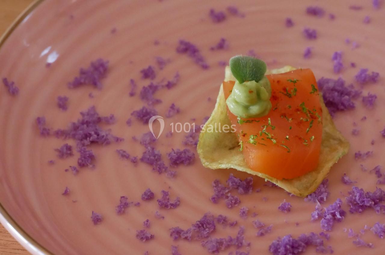Une bouchée de saumon sur une chips, garnie de crème verte, présentée sur une assiette rose avec des éclats violets.