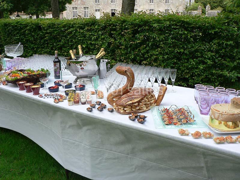 Table dressée en extérieur avec verres, bouteilles, pains, amuse-bouches et desserts sur une nappe blanche.