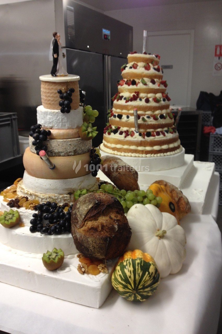 Plateau de fromages en pyramide et gâteau de mariage décorés de fruits, légumes et figurine, dans une cuisine.