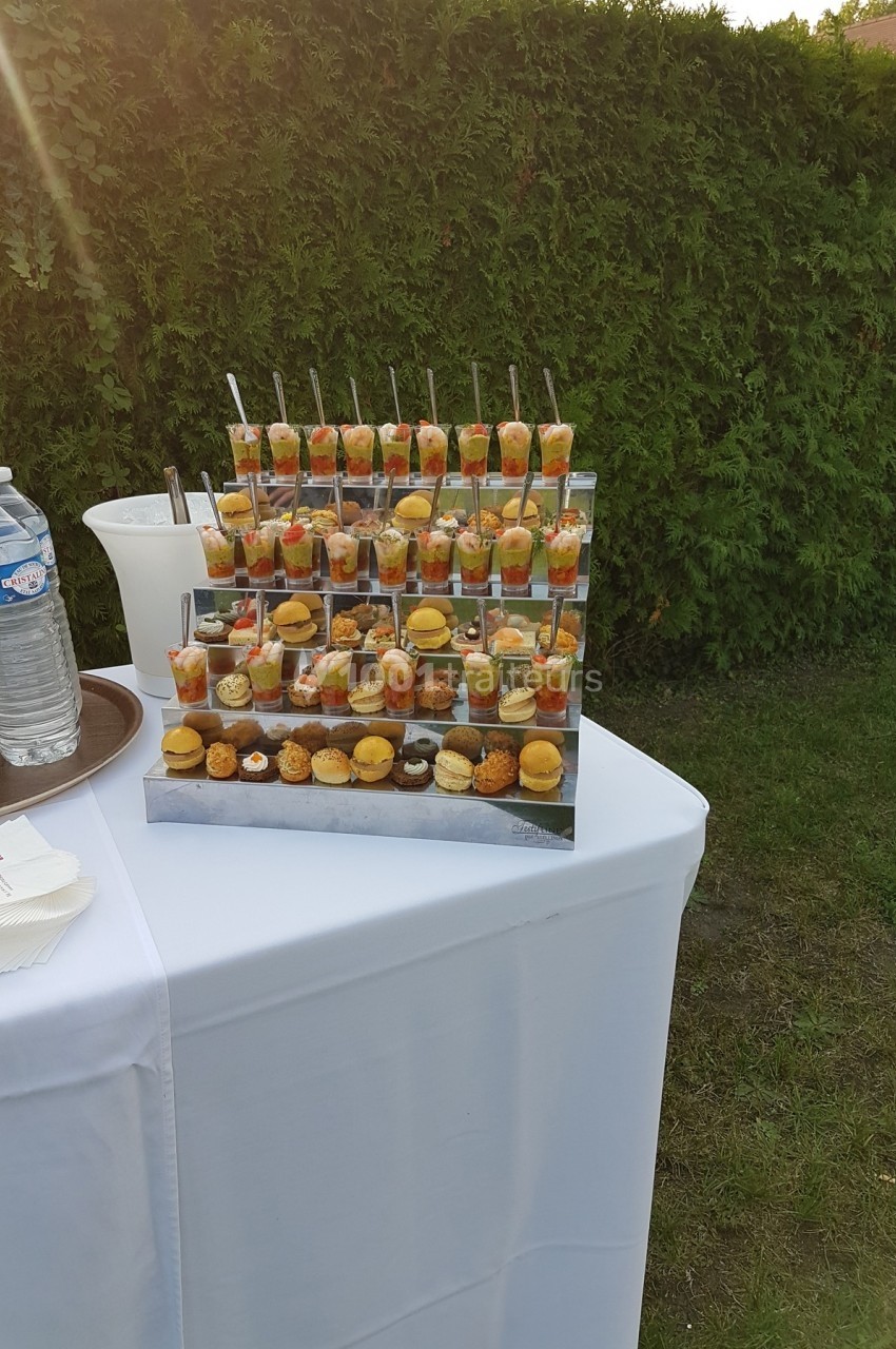 Présentoir de verrines et amuse-bouches variés disposé sur une table blanche en extérieur, près d'une haie verte.