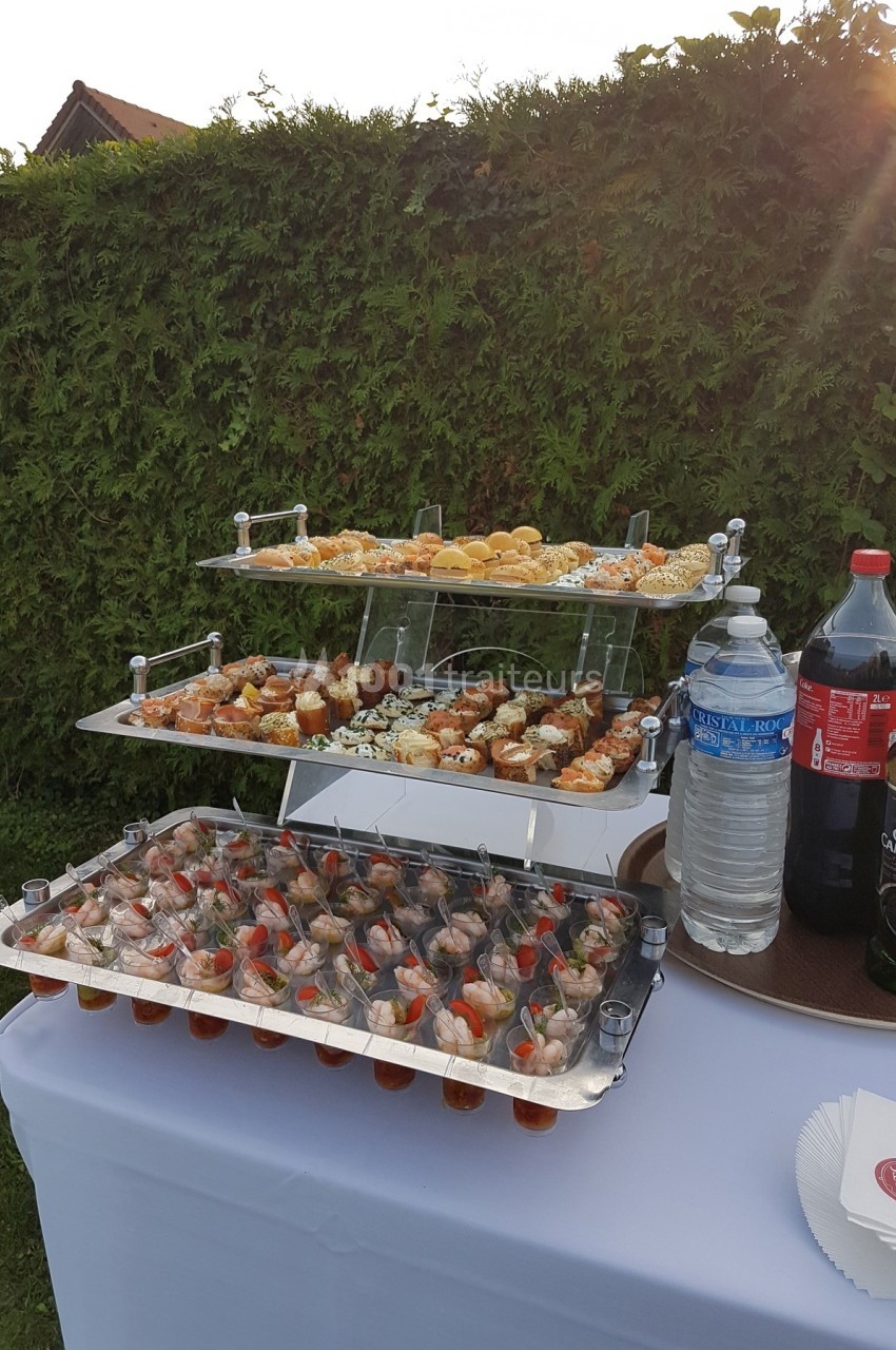 Plateau de buffet avec verrines, amuse-bouches variés et boissons, disposé sur une table en extérieur près d'une haie.