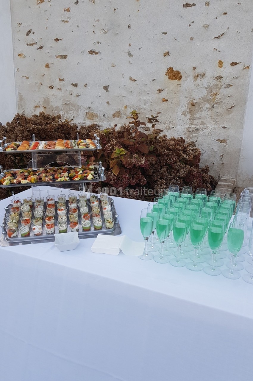 Buffet avec verrines colorées, plateau de fruits et verres de cocktail vert sur une nappe blanche devant un mur en pierre.