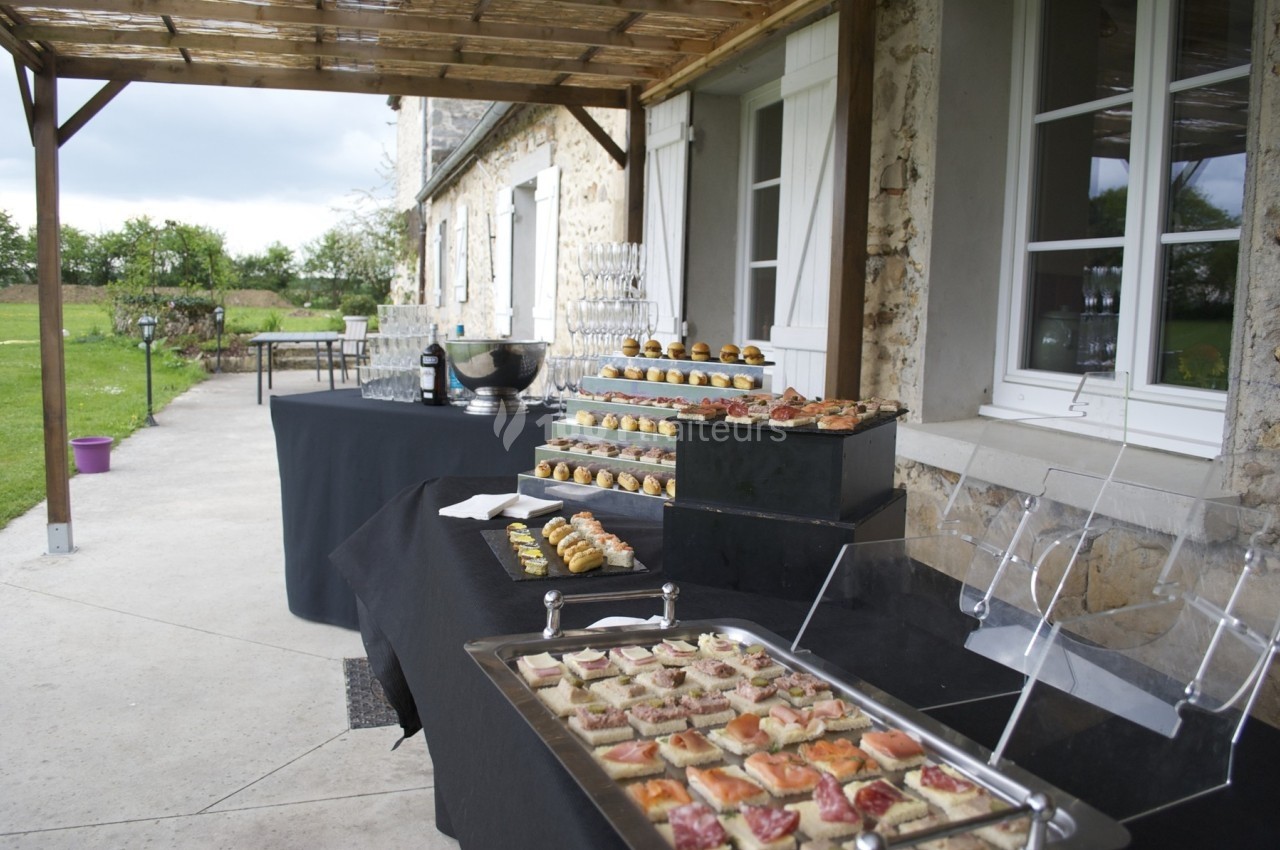 Buffet en extérieur sous une pergola, avec plateaux de canapés, verrines et desserts disposés sur des nappes noires.