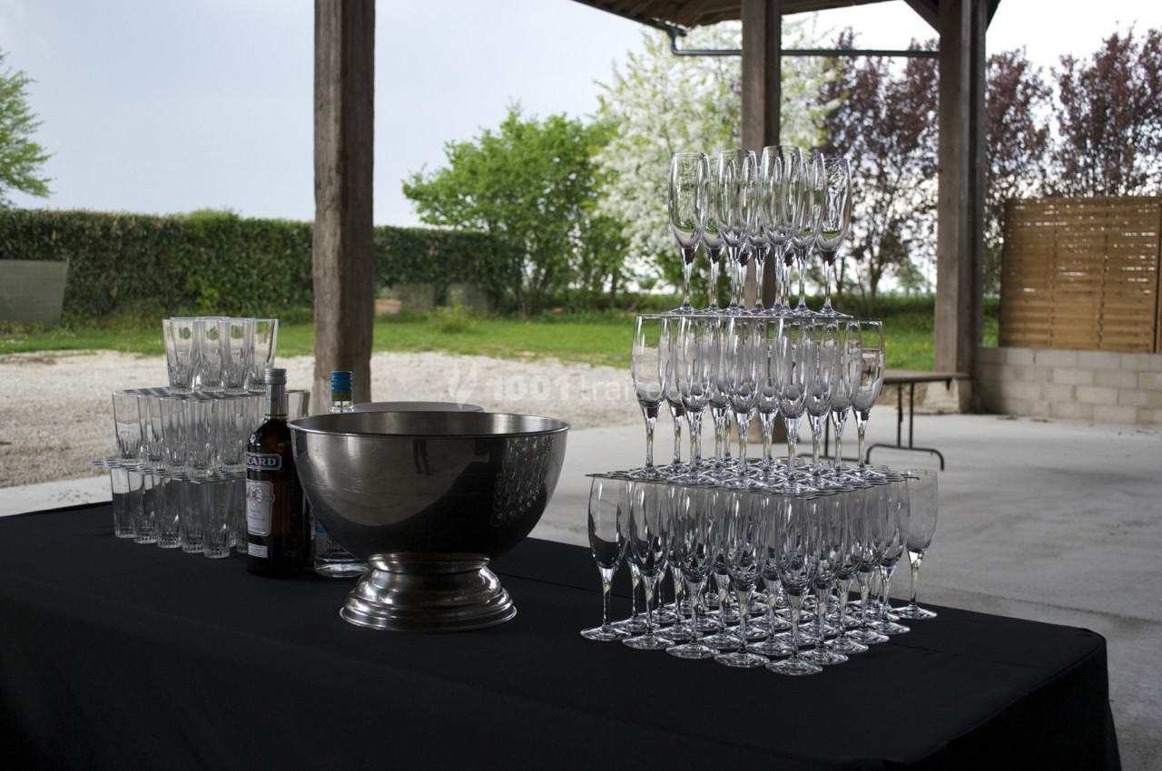 Table avec une nappe noire, des verres empilés en pyramide, une grande coupe métallique et des bouteilles en extérieur.