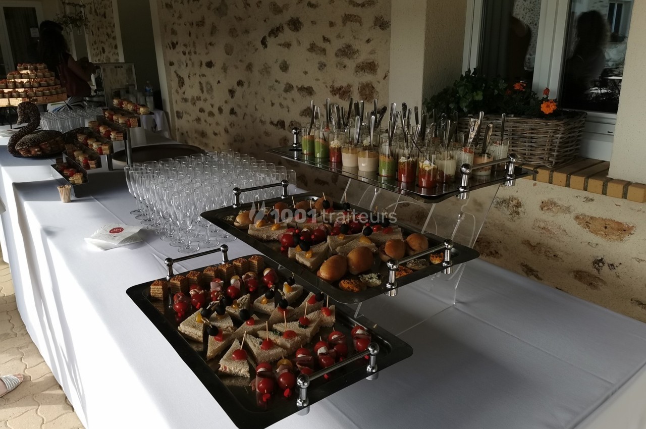 Buffet avec verrines, amuse-bouches variés et verres disposés sur une table blanche en extérieur.