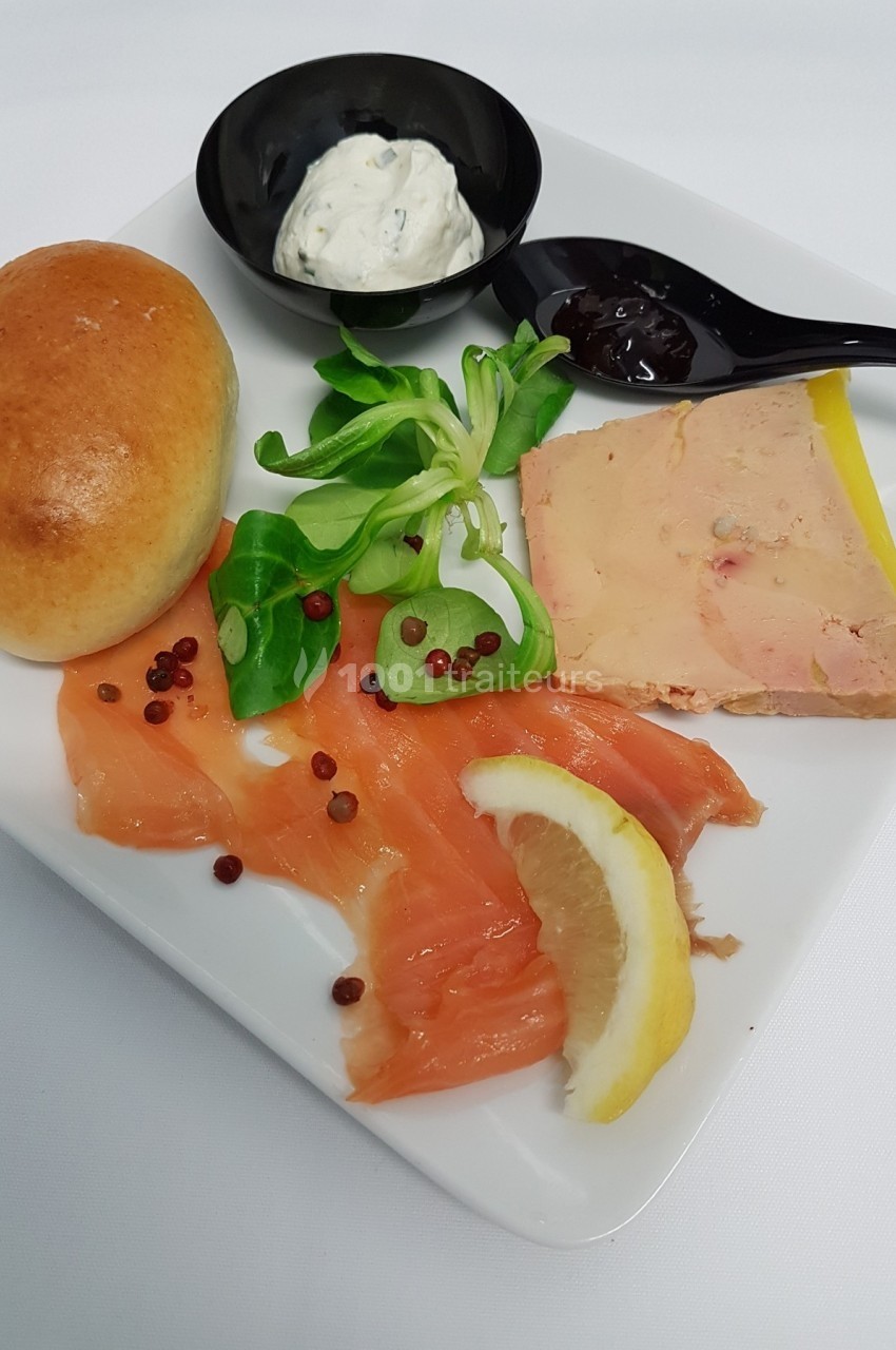 Assiette composée de saumon fumé, foie gras, salade, pain, crème et confiture, présentée sur un fond blanc.