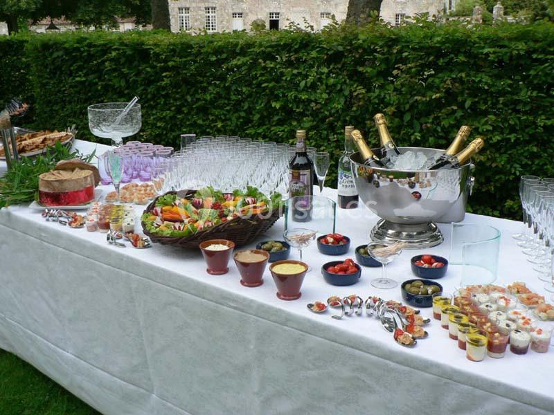 Table de buffet en extérieur avec plats variés, desserts, fruits, bouteilles de champagne et verres alignés.