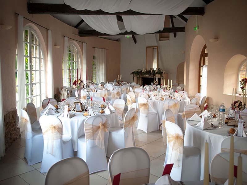 Salle de réception décorée avec des tables rondes, nappes blanches, chaises ornées de nœuds dorés et lumière naturelle.