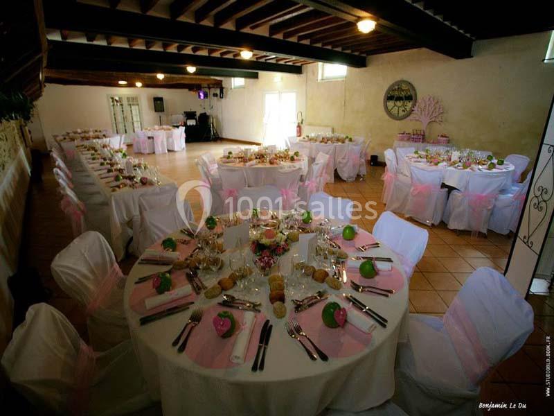 Salle de réception décorée avec des tables rondes dressées, nappes blanches et touches de rose, sous un plafond en poutres…