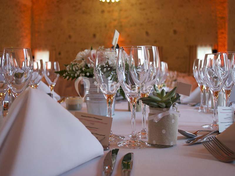 Table dressée avec nappes blanches, verres à vin, serviettes pliées, fleurs et une petite plante en pot.