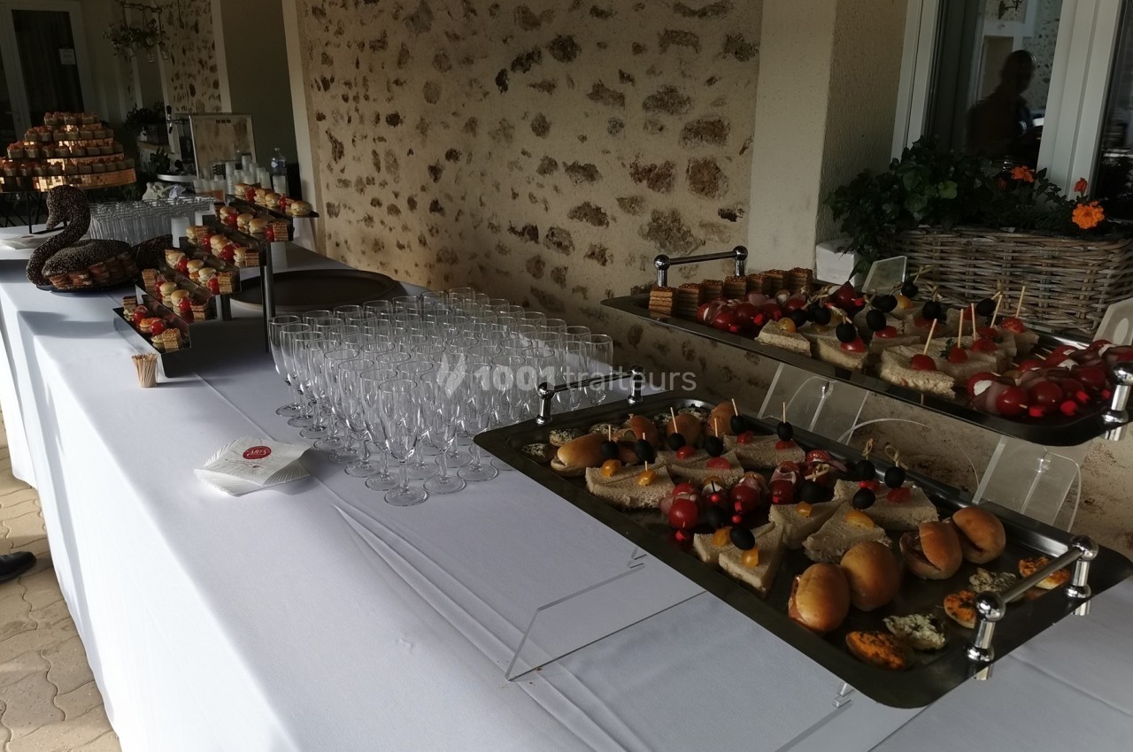 Buffet avec verrines, amuse-bouches, pains garnis et verres disposés sur une table blanche en extérieur.