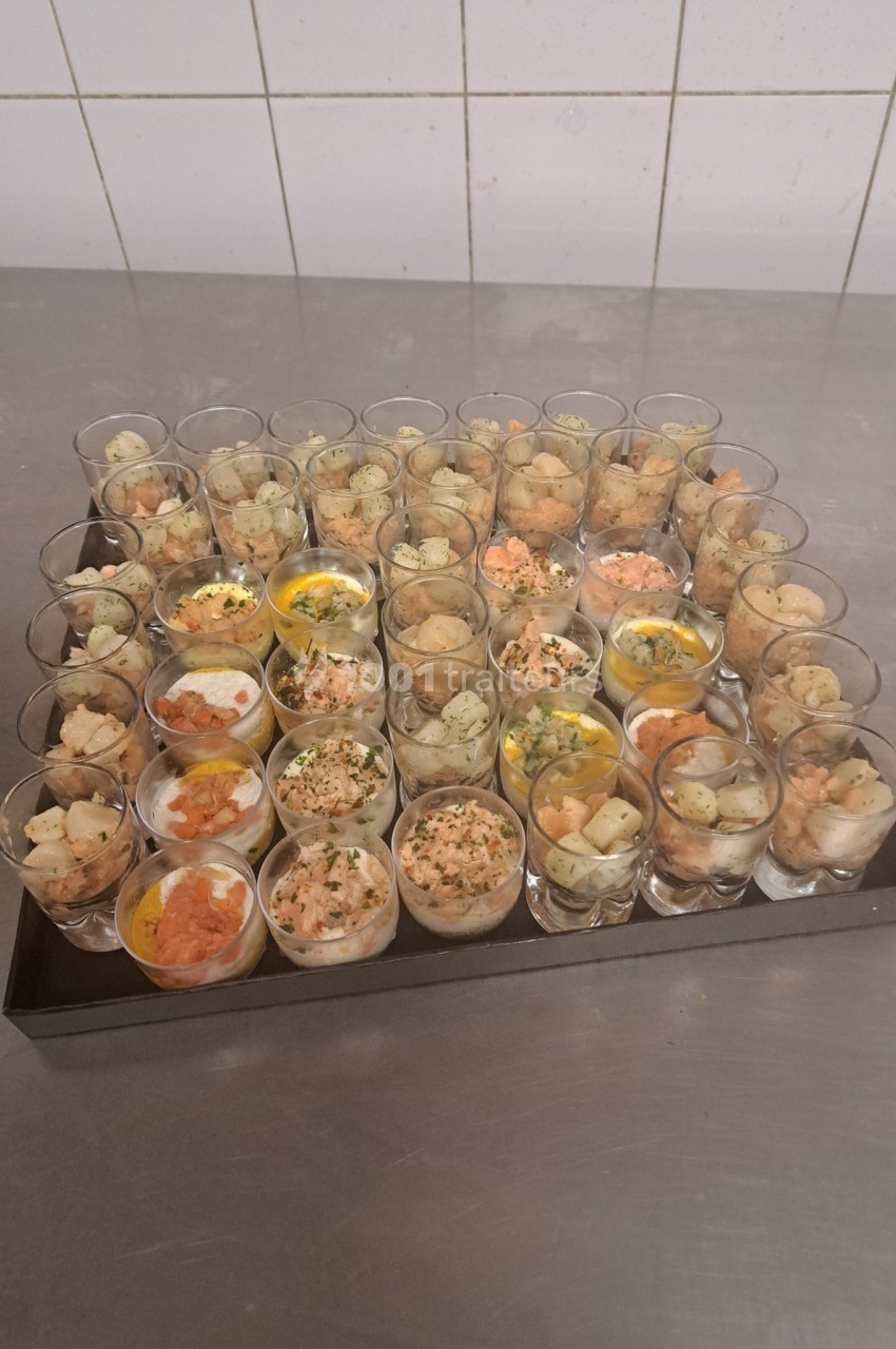 Plateau de verrines variées contenant des ingrédients comme du saumon, des œufs et des herbes, posé sur une table en inox.