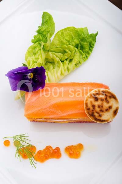 Tranche de saumon fumé avec œufs de poisson, crêpe, feuille de salade et fleur comestible sur assiette blanche.