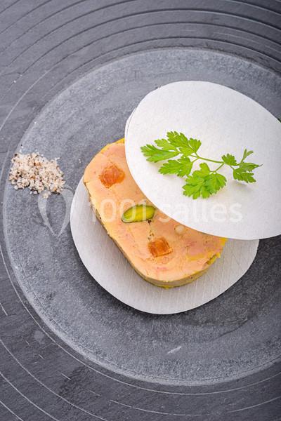 Tranche de foie gras garnie de pistaches et gelée, présentée sur une assiette avec sel et feuille de cerfeuil.
