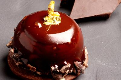 Dessert au chocolat brillant orné de feuille d'or, posé sur une base avec éclats de cacao, sur fond sombre.