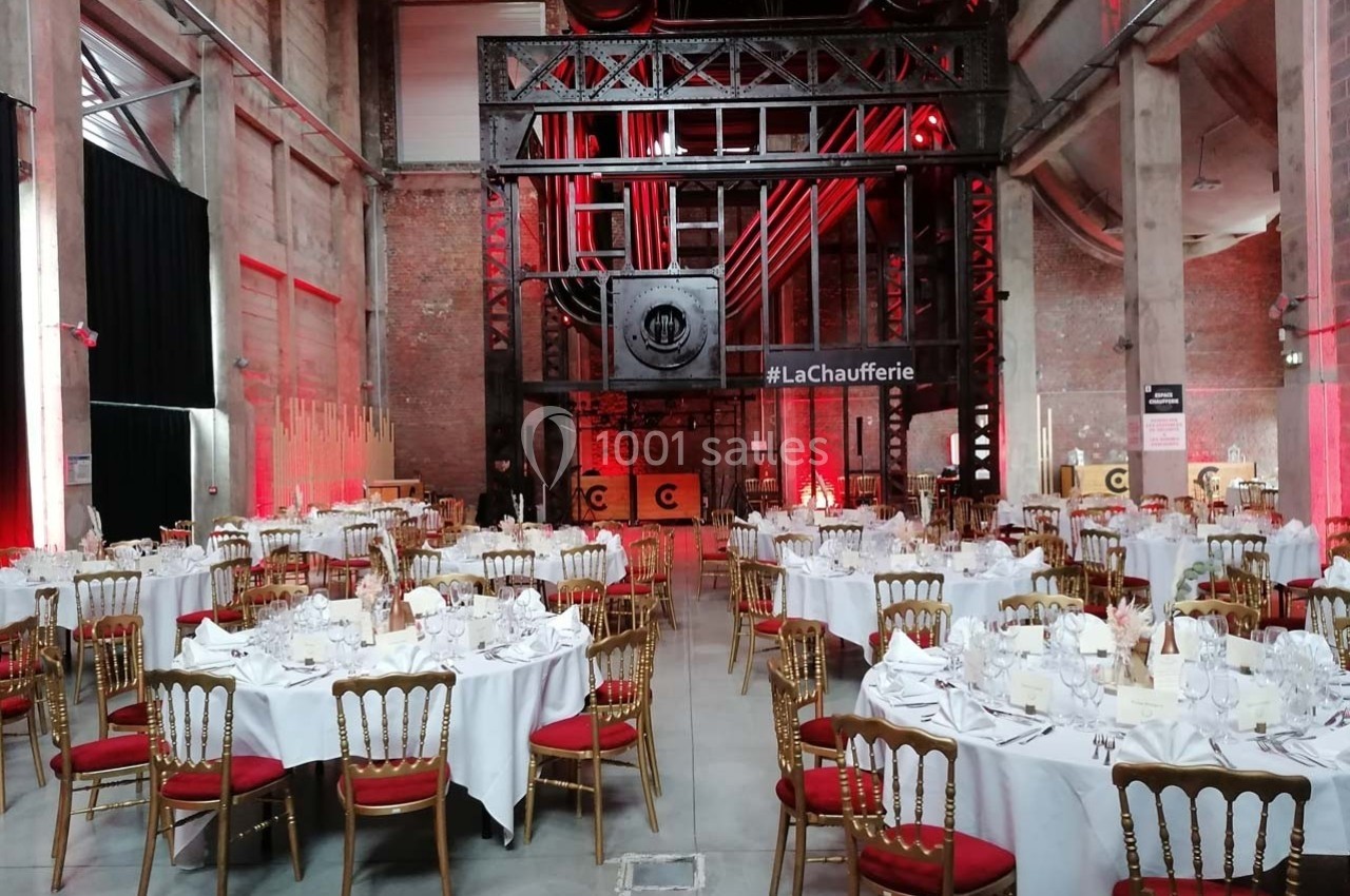 Salle industrielle aménagée pour un événement, avec des tables rondes dressées et des chaises dorées.