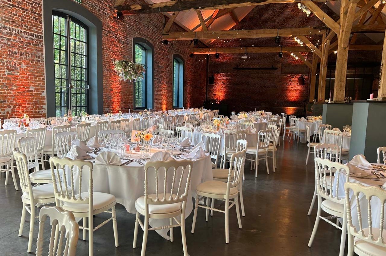 Salle de réception avec murs en briques, grandes fenêtres et tables rondes dressées pour un événement.