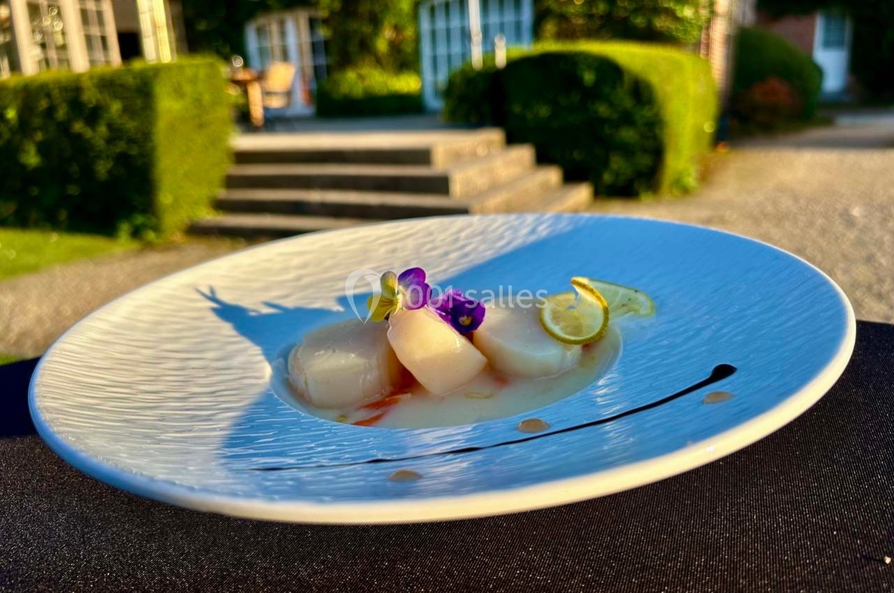 Assiette de coquilles Saint-Jacques décorées de fleurs comestibles et de citron, présentée en extérieur sur une table.