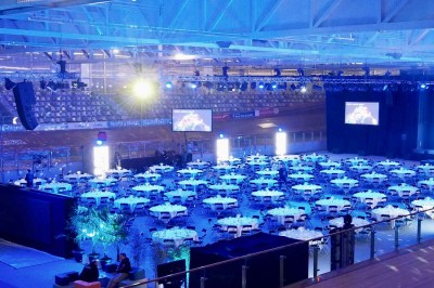 Salle de réception éclairée en bleu avec des tables rondes dressées et des écrans disposés autour.