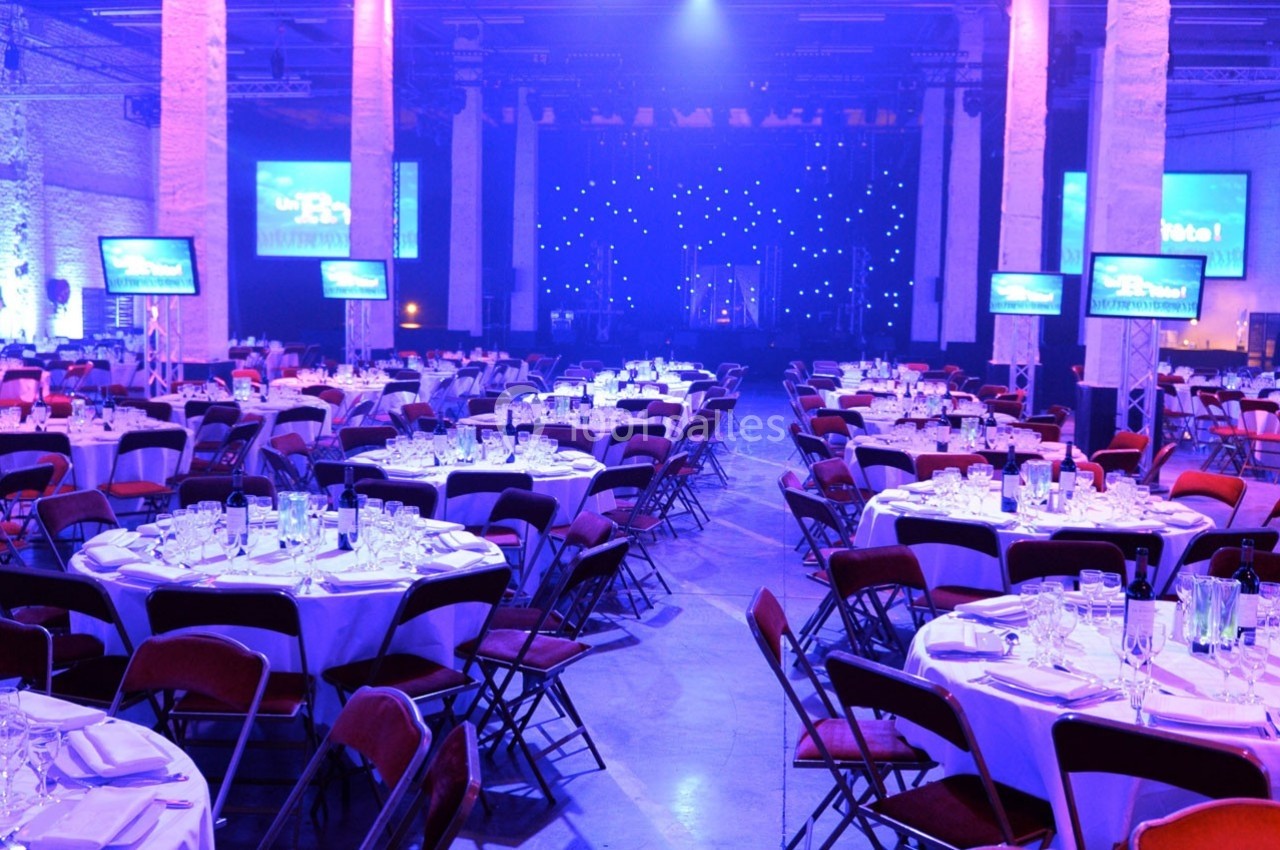 Salle de réception éclairée en bleu avec des tables rondes dressées et des écrans disposés autour.