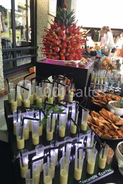 Buffet avec verrines de soupe, brochettes de fruits sur un ananas et assortiments de pains et amuse-bouches.