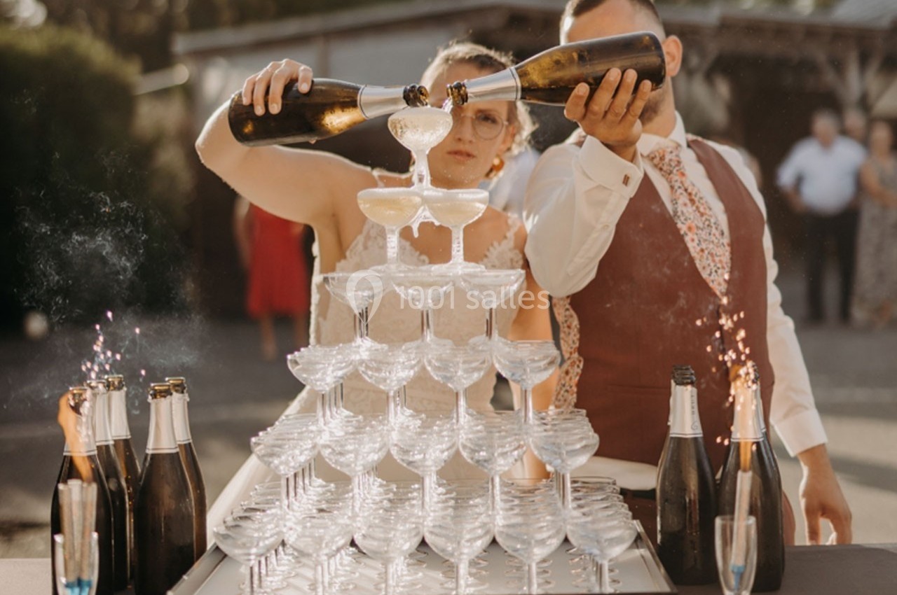 Un couple verse du champagne dans une pyramide de coupes en extérieur, entouré de bouteilles et d'invités en arrière-plan.