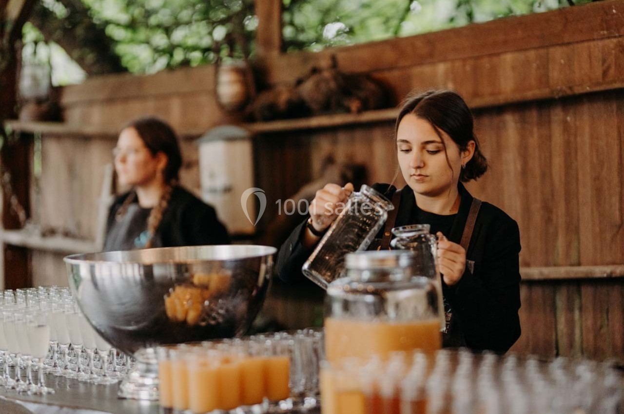 Une femme verse une boisson dans un verre sur un comptoir, entourée de carafes et de verres alignés.
