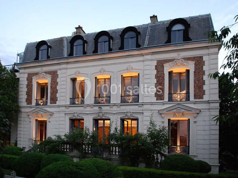 Façade d'une maison bourgeoise à trois étages, éclairée de l'intérieur, entourée de végétation.