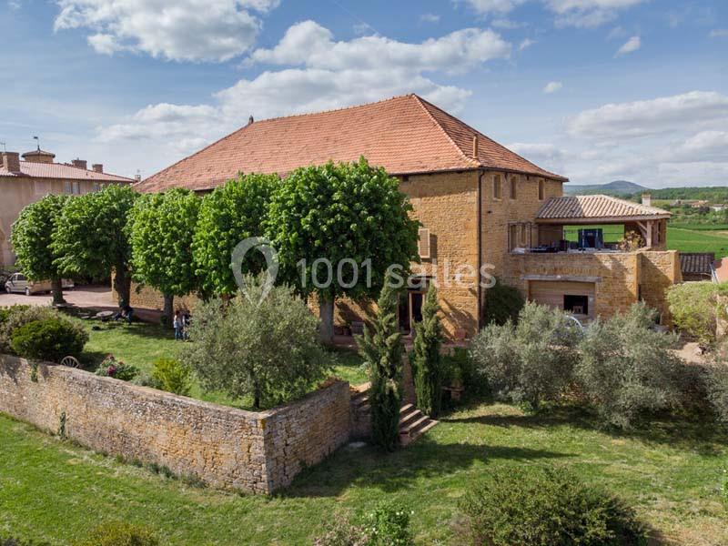 Grande maison en pierre avec toit en tuiles rouges, entourée d'arbres et d'un jardin verdoyant sous un ciel partiellement…
