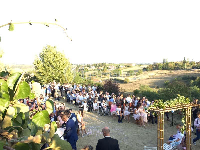 Un mariage en plein air avec des invités assis, un paysage rural en arrière-plan et une arche décorative au premier plan.