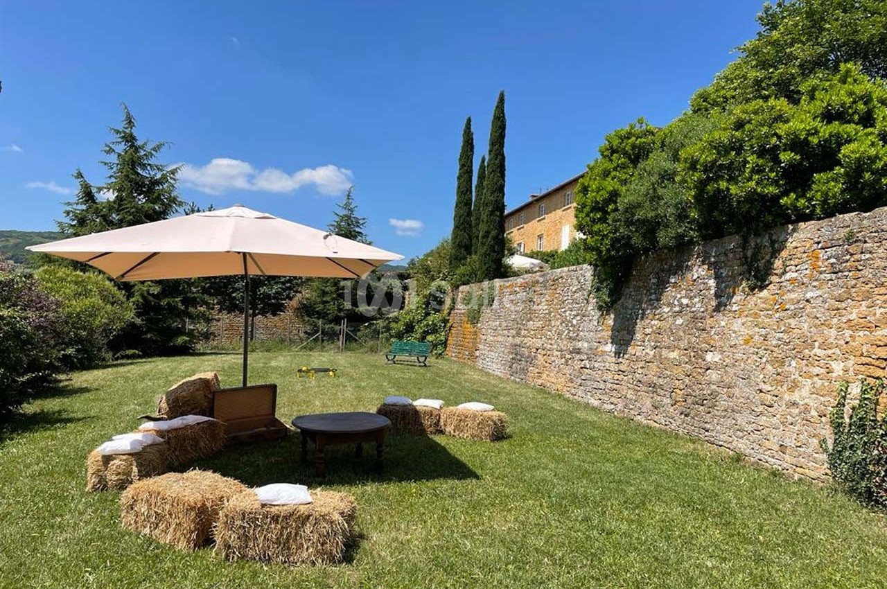 Espace extérieur avec parasol, bottes de paille en sièges, table basse, pelouse et mur en pierre sous un ciel bleu.