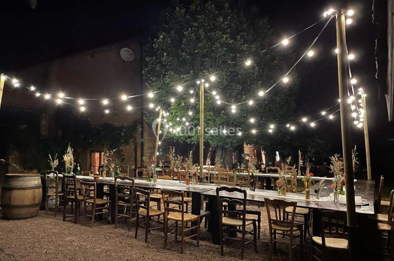 Tables en bois décorées de fleurs et chaises dépareillées sous des guirlandes lumineuses dans une cour extérieure la nuit.