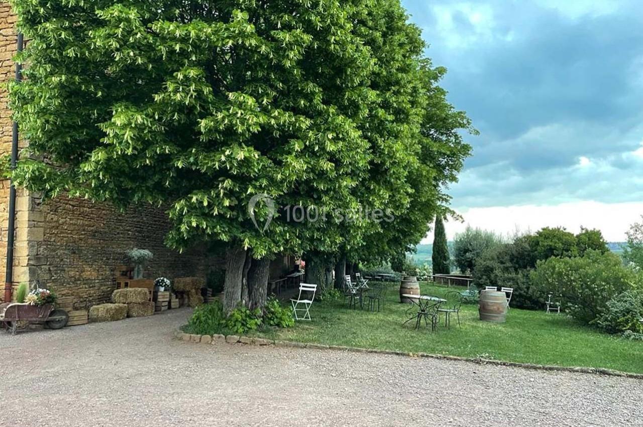 Espace extérieur avec des arbres, des tables et chaises en métal, sur fond de pelouse et paysage rural.