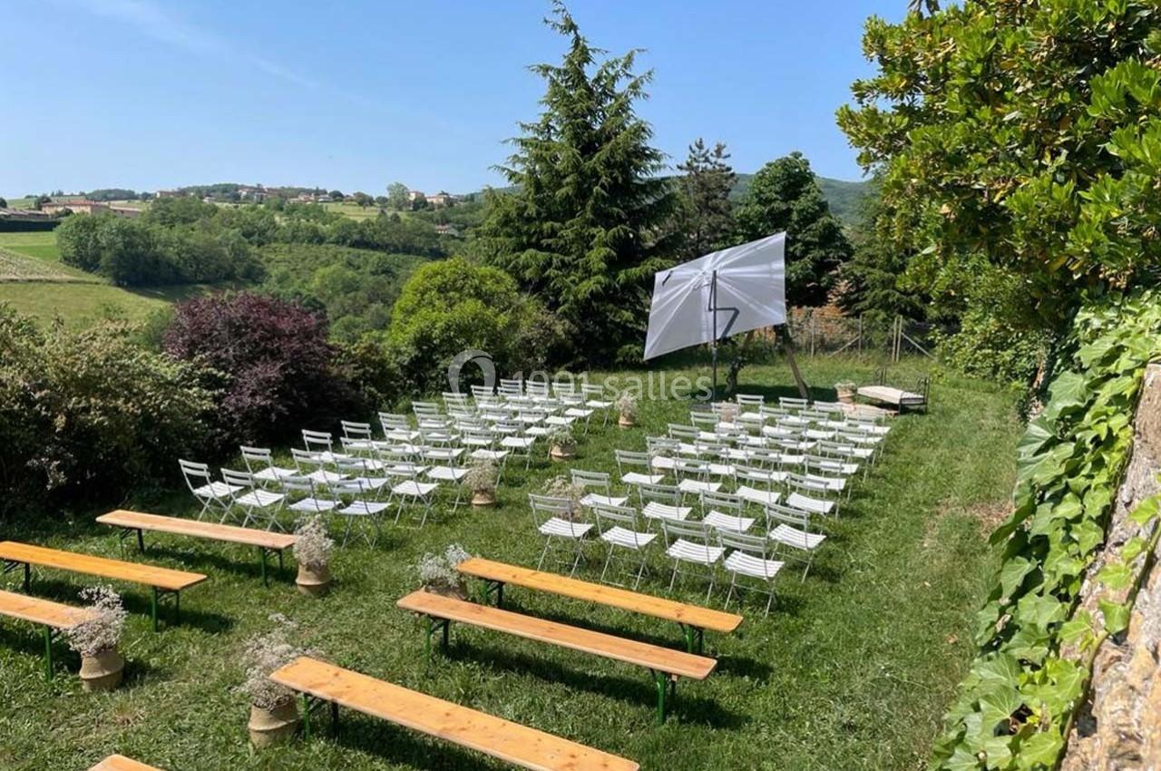 Chaises blanches et bancs en bois disposés en extérieur, face à un écran, entourés de verdure et d'arbres.