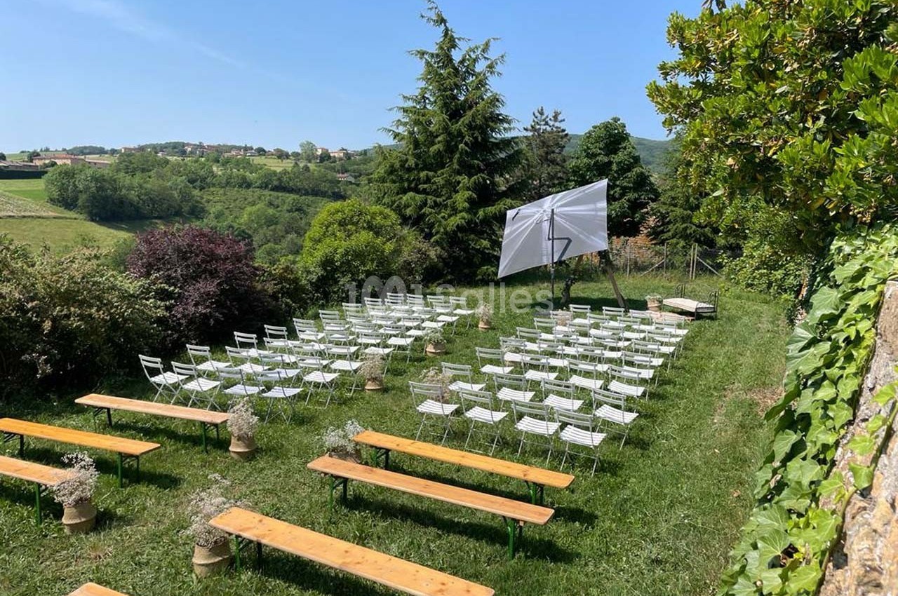 Chaises blanches et bancs en bois disposés en extérieur, face à un écran, entourés de verdure et d'arbres.