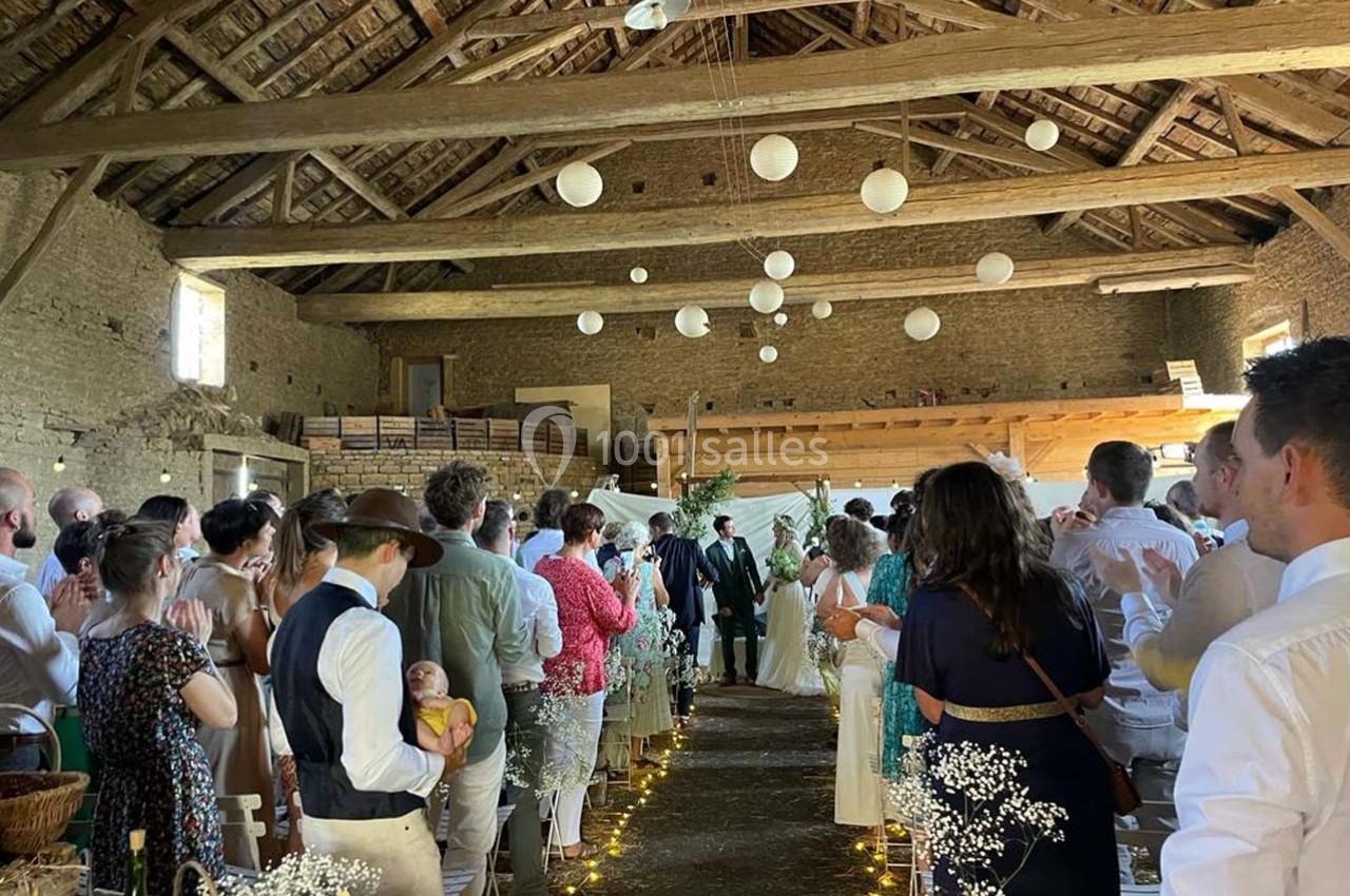 Cérémonie de mariage dans une grange en pierre, avec invités assis et décorations lumineuses suspendues.
