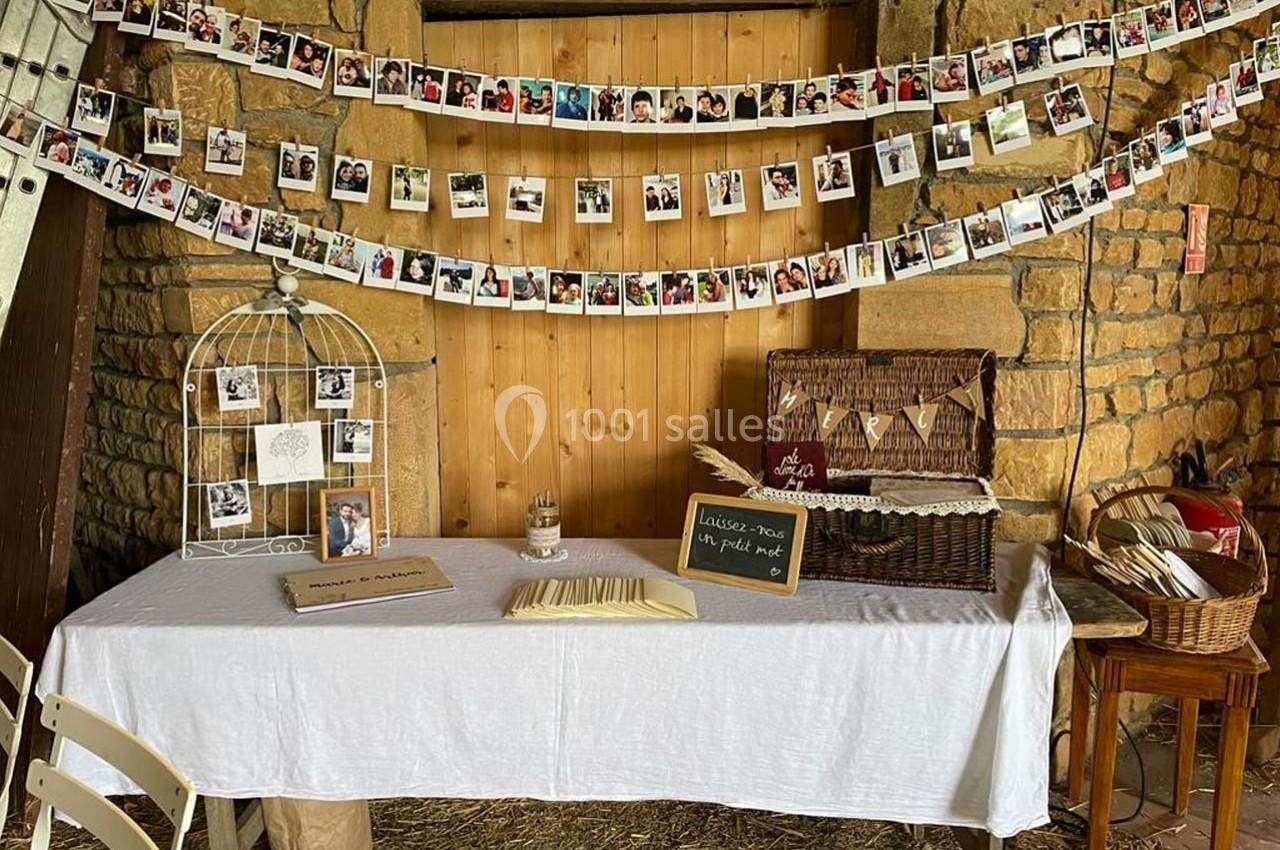 Table décorée avec nappe blanche, photos suspendues, cage décorative, panier en osier et livre d'or dans une grange.