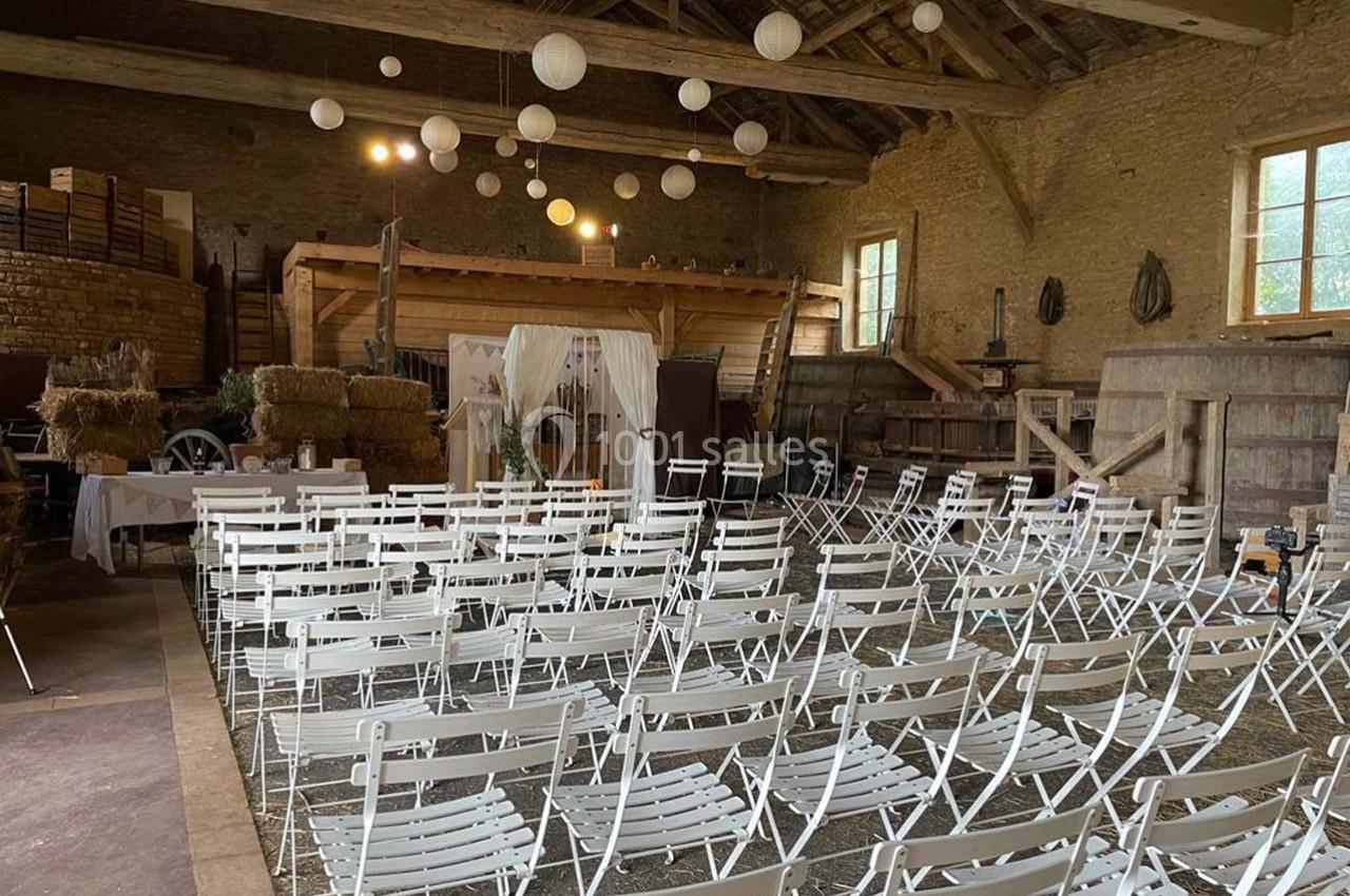 Salle rustique aménagée avec des chaises blanches alignées, décorations suspendues et éléments en bois et paille.