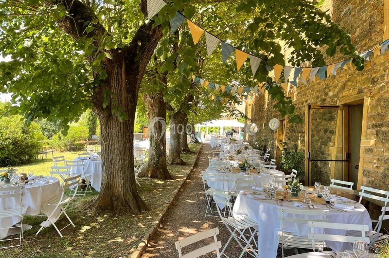 Tables décorées pour un repas en plein air sous des arbres, avec guirlandes suspendues et bâtiment en pierre en arrière-plan.