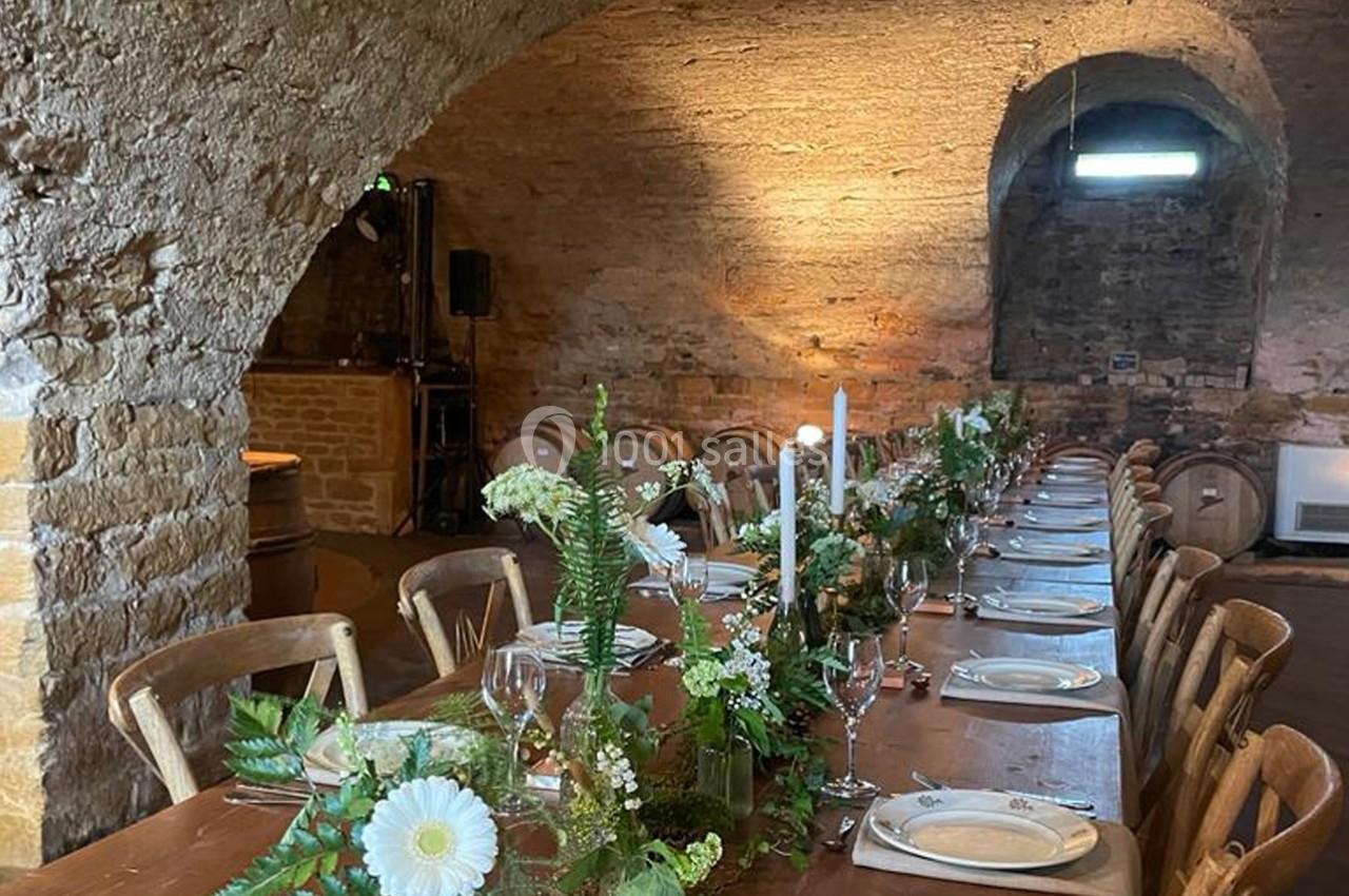 Table en bois décorée de fleurs et bougies dans une cave voûtée en pierre avec des tonneaux en arrière-plan.