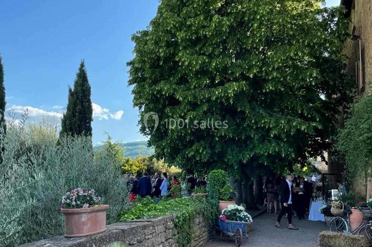 Un chemin bordé de végétation mène à un rassemblement sous un grand arbre, avec des pots de fleurs et une charrette…