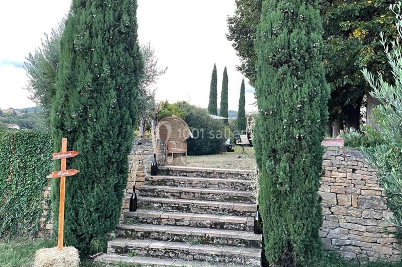 Escalier en pierre bordé de cyprès menant à un jardin avec fauteuil en osier, panneaux directionnels sur botte de foin.