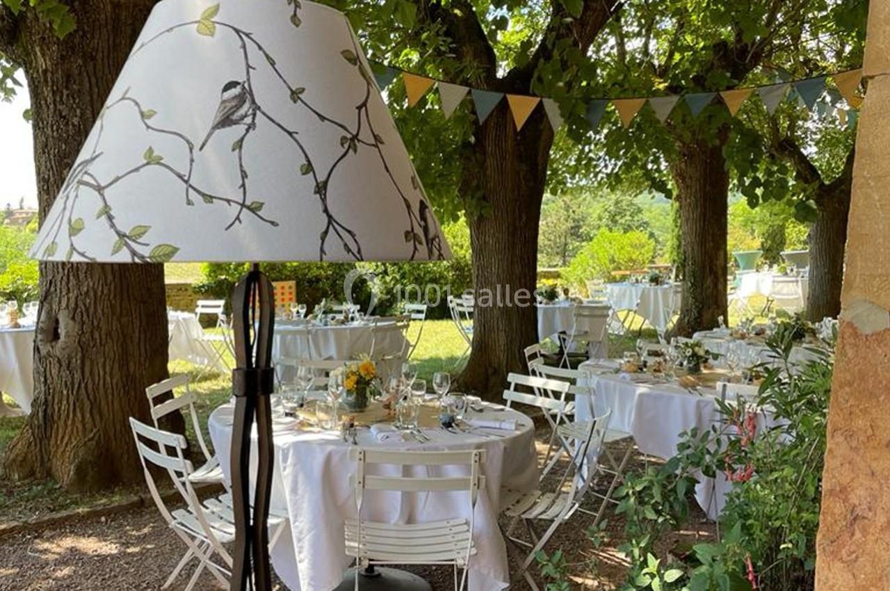 Lampadaire décoratif devant des tables rondes dressées sous des arbres, dans un jardin ombragé avec guirlandes suspendues.