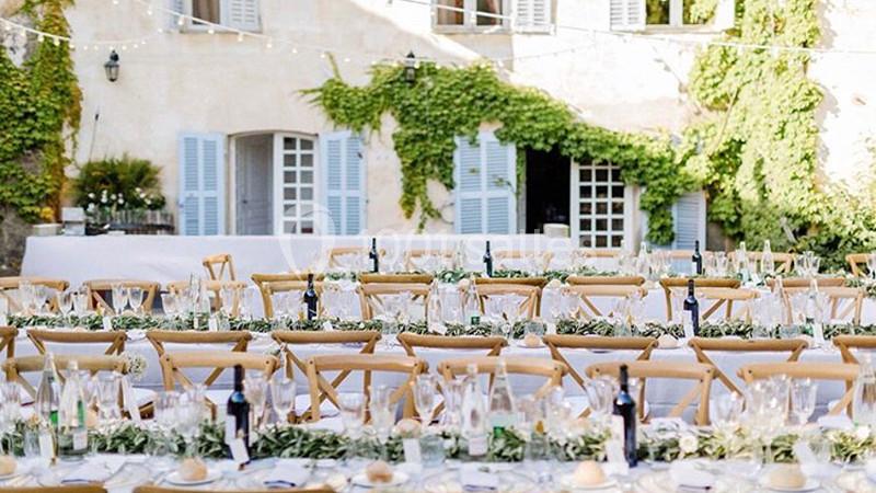 Tables décorées pour un événement en plein air, disposées devant une maison aux volets bleus et murs couverts de lierre.