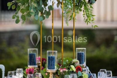 Tables décorées pour un événement en plein air, disposées devant une maison aux volets bleus et murs couverts de lierre.