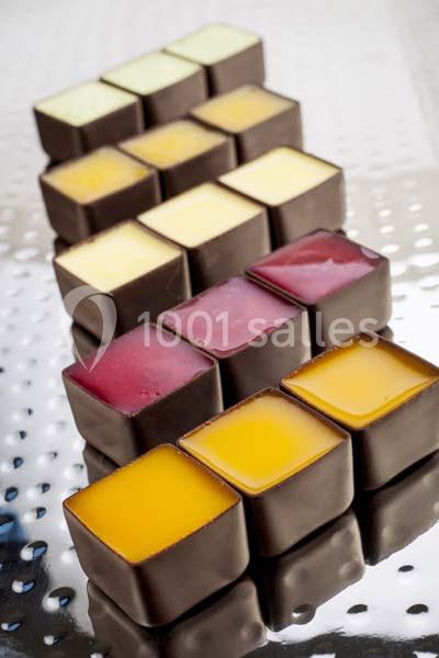Chocolats carrés garnis de différentes crèmes colorées, disposés en rangées sur une surface métallique réfléchissante.