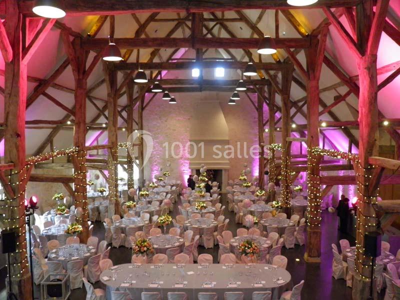 Grande salle de réception avec charpente en bois, tables rondes dressées et décorations lumineuses.