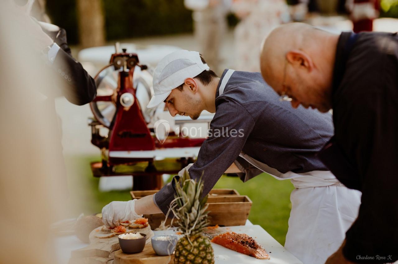 Un cuisinier prépare des plats avec des ingrédients frais sur une table en extérieur, devant une trancheuse rouge.