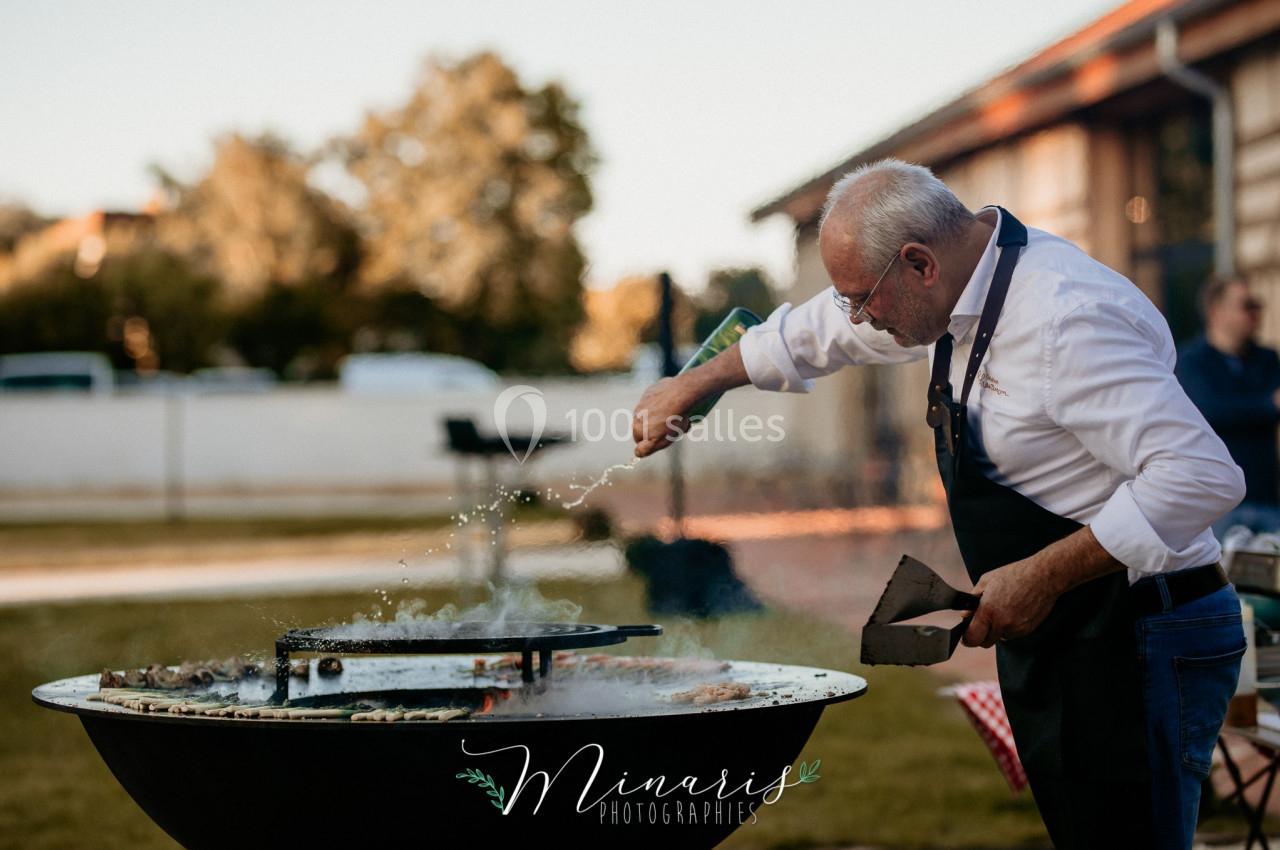 Un homme en tablier cuisine sur un brasero en plein air, saupoudrant des ingrédients sur une plaque chauffante.