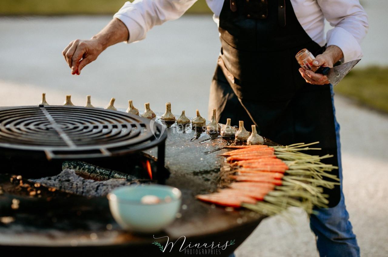 Une personne assaisonne des légumes grillés, dont des carottes et des artichauts, sur une plancha en plein air.